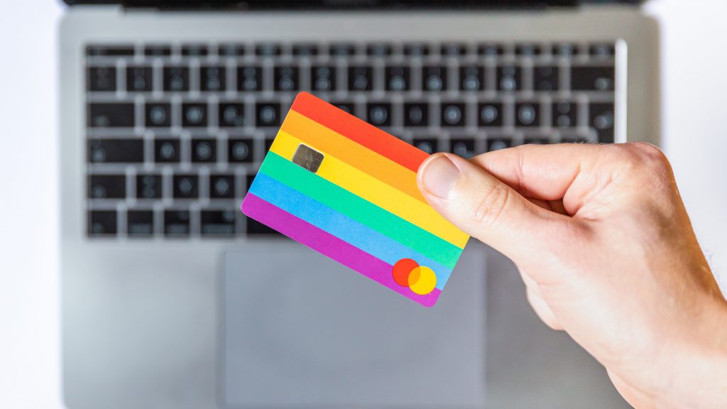 Mão segurando cartão de crédito com estampa de arco-iris, sobre teclado de notebook.