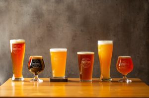 vários copos espececíficos para apreciar cada tipo de cerveja, cheios de cervejas, sobre uma mesa de madeira envernizada. Ao fundo uma parede de cimento queimado.