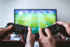 Duas mãos segurando dois controles de vídeo game lado a lado, em frente a uma televisão, onde um jogo de futebol aparece.