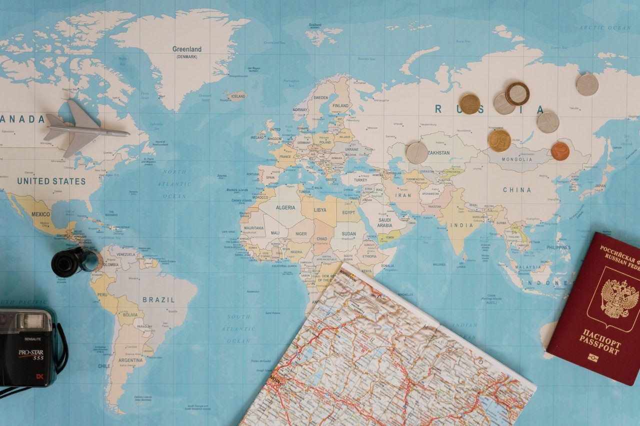 Há um mapa do mundo aberto com algumas moedas e um avião de brinquedo na parte superior, enquanto na parte inferior há uma câmera, passaporte e um mapa fechado.