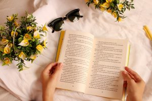 Na imagem há uma pessoa segurando um livro com as duas mãos sobre uma superfície branca. Próximo ao livro há dois arranjos de flores amarelas e um óculos de sol.