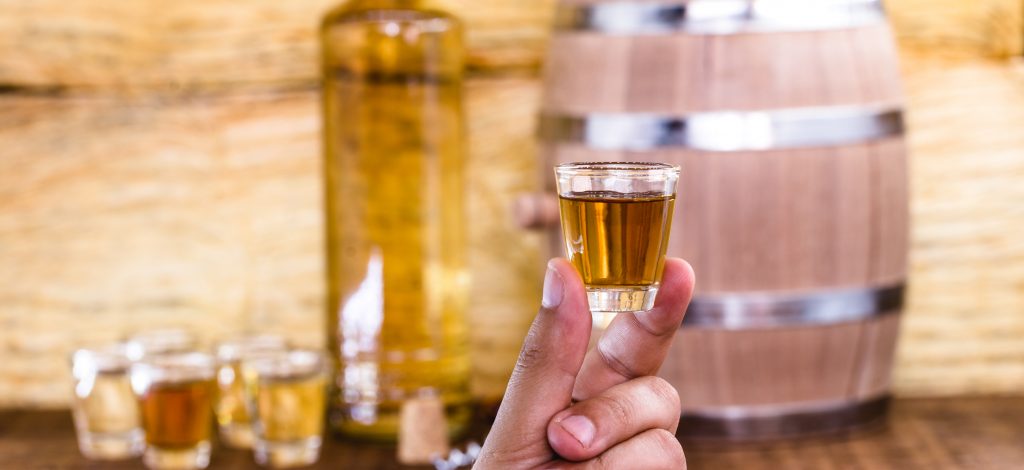 Na imagem há alguém segurando um copo pequeno com um líquido de cor dourada. Ao fundo há um barril de madeira com uma garrafa de vidro ao lado contendo um líquido de cor dourada e alguns copos próximos.