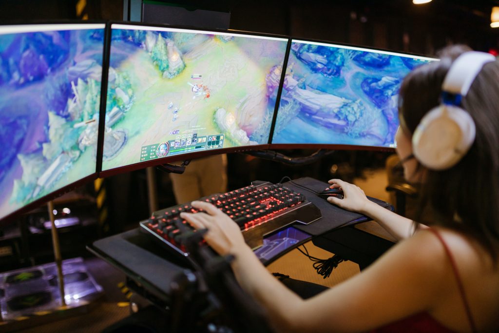 Na imagem há uma pessoa em frente a três telas de computador reproduzindo o jogo League of Legends, sendo que a pessoa está com a mão direita mexendo no mouse e com a mão esquerda digitando no teclado.