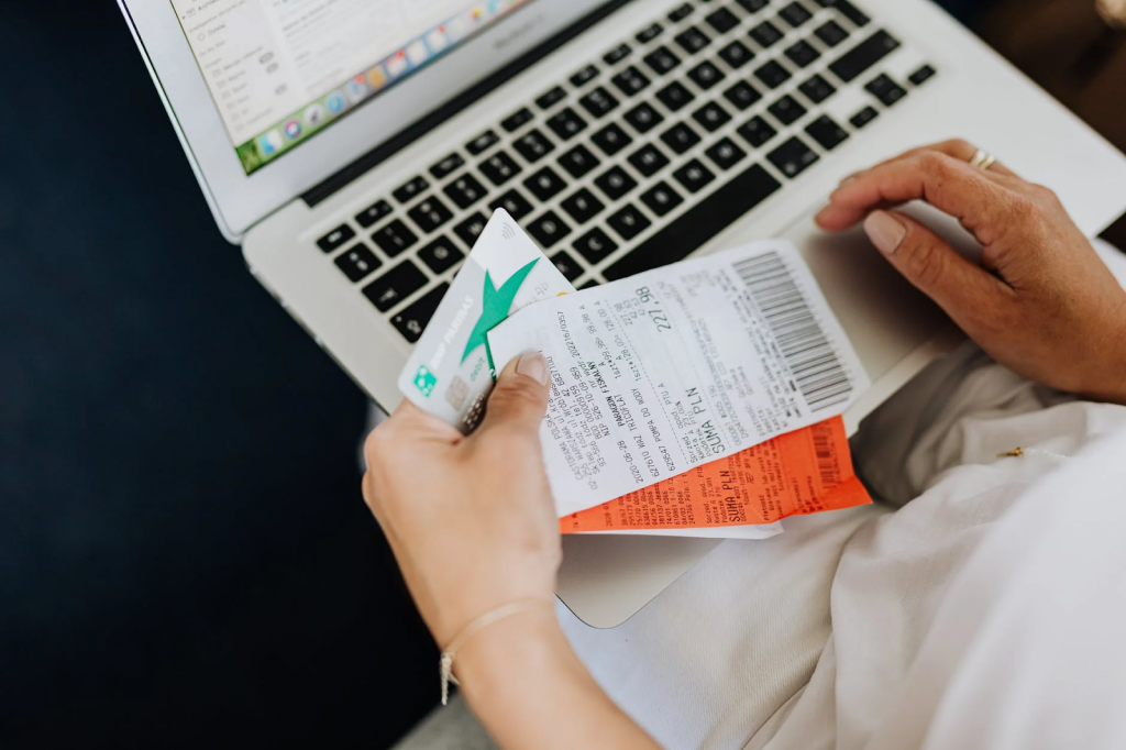 Mulher de roupa branca segurando um cartão de crédito e algumas notas fiscais, mexendo em um notebook aberto em uma superfície azul.