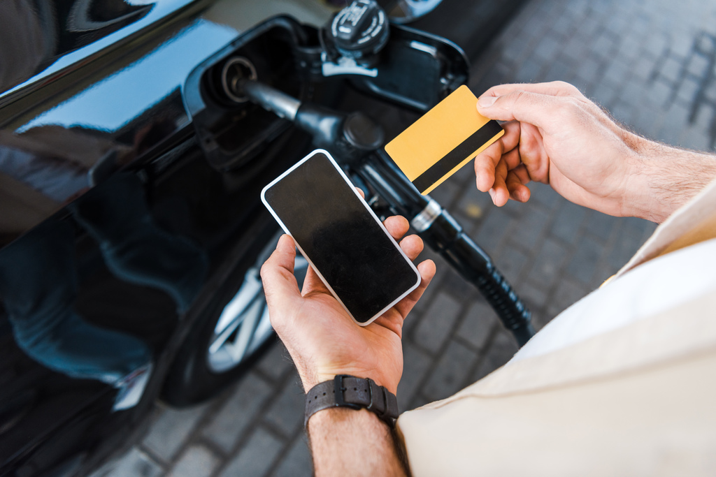 Homem abastecendo seu carro e segurando um smartphone e um cartão de crédito.