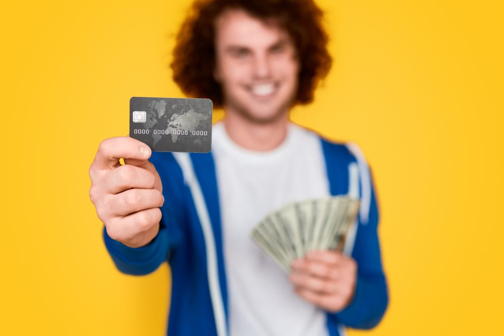 Homem de cabelos enrolados vestindo uma camisa branca e uma jaqueta azul, ele está esticando a mão esquerda para frente e mostrando um cartão de crédito, enquanto segura cédulas de dinheiro com a direita rente ao corpo.
