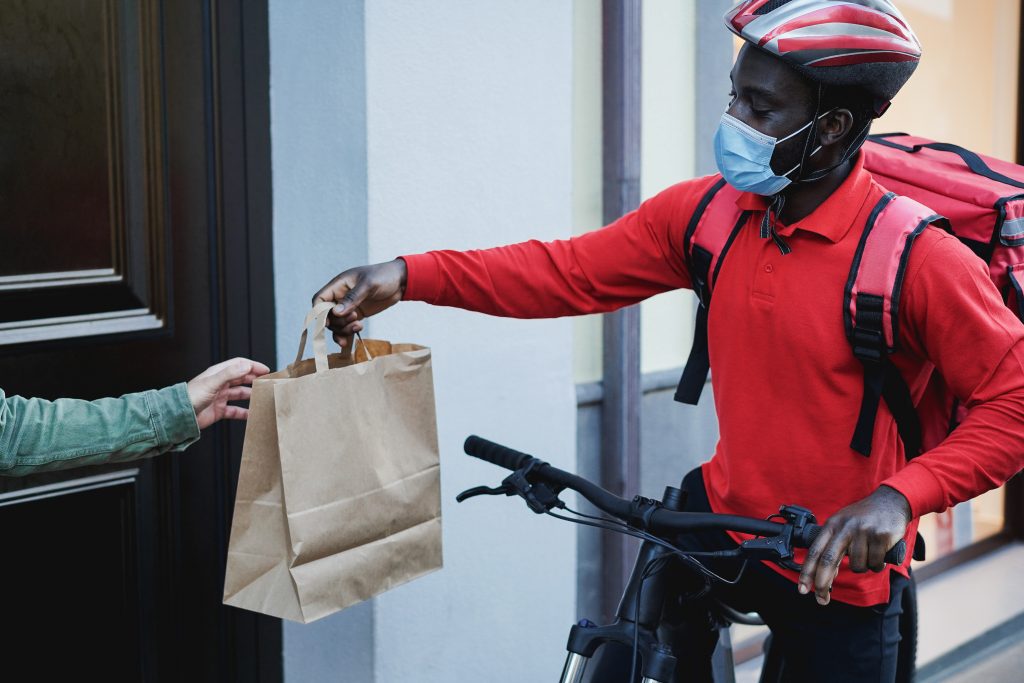 Imagem de uma porta de casa do lado de fora, do lado esquerdo há uma mão pegando um pacote de entrega e, do lado direito, há um homem em uma bicicleta com uma mochila nas costas entregando esse pacote. 