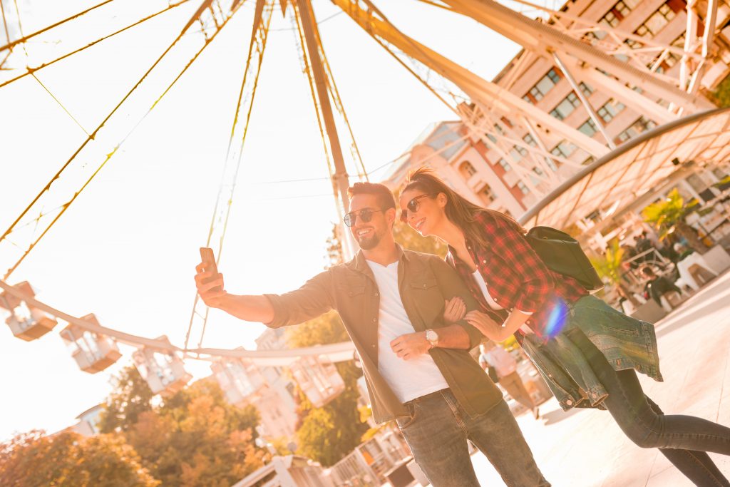 Imagem de um parque de diversões com uma roda gigante ao fundo e na frente um casal tirando foto, um homem alto fazendo a selfie e do lado direito dele uma mulher. 